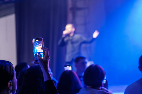 Crowd watches live singer on stage with phone capturing moment amid blue lights creating vibrant concert atmosphere - Powered by Adobe