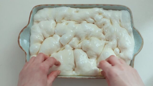 Close-up of raw focaccia dough in baking tray being massaged with hands, glistening with olive oil and forming air bubbles