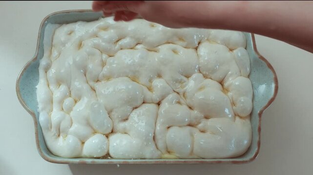 Hand sprinkling coarse salt over raw focaccia dough in ceramic baking dish, glossy surface with air bubbles forming from fermentation, preparation of traditional homemade bread