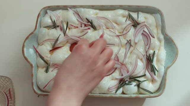 Hand placing cherry tomatoes onto raw focaccia dough in ceramic baking dish, already topped with red onion and rosemary, oily surface full of bubbles during final decorative stage before baking