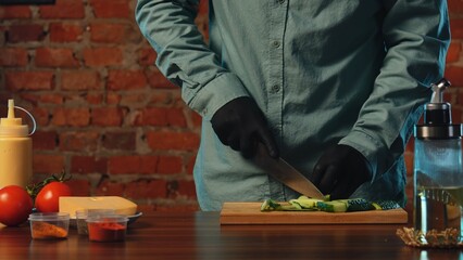 A chef wearing black gloves slices fresh cucumbers on a wooden board in a rustic kitchen setting. The process of preparing food in a cafe or restaurant.
