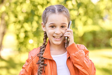 Caucasian young girl in orange jacket talking on smartphone in park with green background.