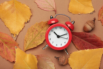 Red alarm clock, acorns and autumn leaves on brown background. Daylight saving time end