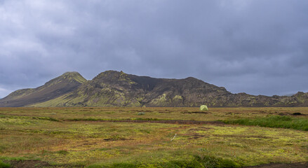 Green tent in remote Icelandic field with mossy hills and dramatic cloudy mountain backdrop.
