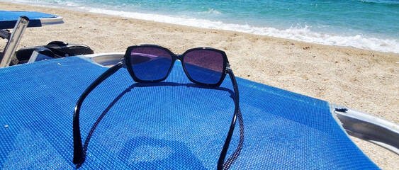 A pair of dark-framed sunglasses rests on a blue beach chair by the sea. A sandy beach and turquoise water with gentle waves are visible in the background. A sunny day, relaxation, and carefree mood.