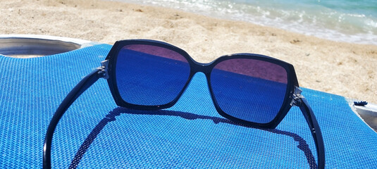 A pair of dark-framed sunglasses rests on a blue beach chair by the sea. A sandy beach and turquoise water with gentle waves are visible in the background. A sunny day, relaxation, and carefree mood.