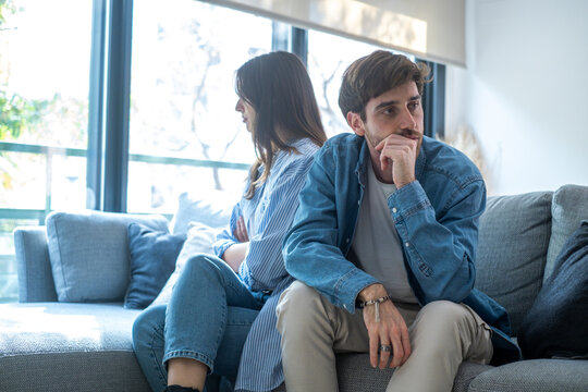 Fototapeta Young couple sitting on sofa at home arguing and confronting each other, concept of relationship problems, breakup, jealousy, betrayal, lack of trust, or communication issues