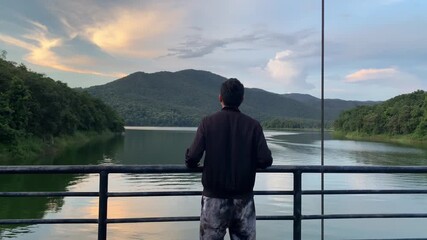 a man standing on a bridge or dam railing, overlooking a calm reservoir or lake surrounded by forested mountains