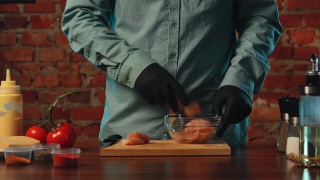A chef seasoning raw chicken with spices. Ingredients include tomatoes, cheese, and condiments on a table.