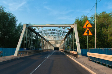 Nowa Sól - Stalowy most kratownicowy rozciąga się nad rzeką Odrą w ciągu drogi nr 315, otoczony niebieskimi barierkami i bujnymi drzewami. Ciepłe światło podkreśla detale konstrukcyjne. © Pol Badger