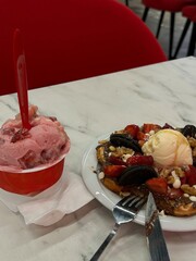 Plate with waffle topped with ice cream, strawberries, and cookies next to a cup of strawberry ice cream on a marble table.