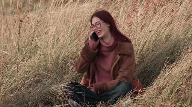 Teen girl smiling while talking on her phone, seated in tall grass, dressed warmly in a brown coat and pink sweater, enjoying an autumn day.