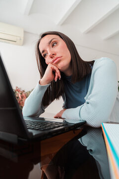 A young woman displays signs of stress and contemplation as she engages with her laptop, reflecting on her academic challenges and the pressures of student life while seated at a table at home