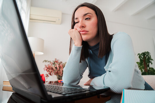 A young woman looking frustrated while working on her laptop at home, showing signs of sadness and stress on her face as she attempts to concentrate on her studies and tackle her academic tasks