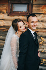 Fototapeta premium Bride in an elegant white gown and groom in a classic suit pose by a rustic wooden wall. She holds a bouquet of white flowers. A warm atmosphere of love, style, and intimate romance.