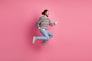 Young bearded man joyfully jumping against a pink backdrop wearing a colorful sweater, exuding happiness and energetic vibes