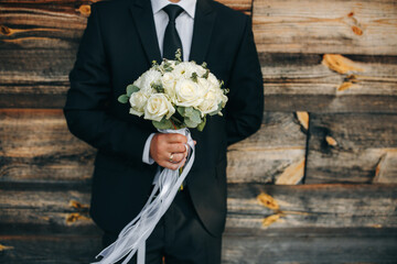 The groom in a classic black suit holds a white rose bouquet against a wooden wall. Stylish wedding detail, elegant contrast of colors, and a romantic atmosphere highlight the special moment.