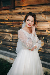 Bride in white dress holding bouquet near rustic wooden wall. Her veil flows in the wind, creating a soft, romantic, countryside atmosphere.