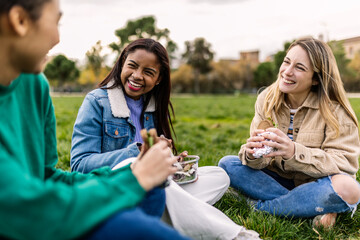 Group of happy young women students laughing while having lunch outdoors on green grass. Friendship and relaxation concept