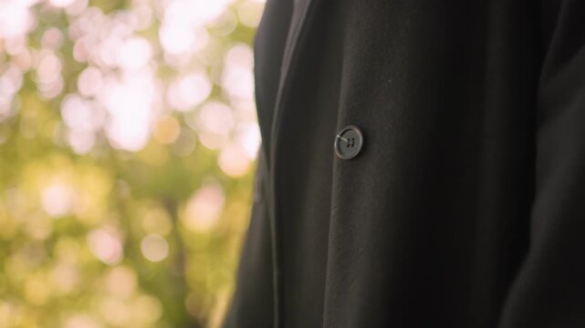 Winter coat with side button worn by female while walking, detail of smooth dark fabric and glossy fastener in focus, gentle motion suggesting cool season stroll under green canopy