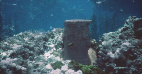 Beneath the surface of clear blue waters, a weathered stone column stands amid coral formations. Fish swim nearby, exploring the mysterious underwater ruins.