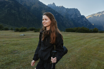 Naklejka premium A young woman in outdoor gear stands in a sunlit grassy field with rugged mountains in the distance, smiling softly as she holds a trekking pole and prepares for adventure.