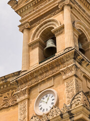 Timeless elegance: Ornate bell tower of a Maltese cathedral, with Roman numeral clock and intricate stone carvings echoing Baroque grandeur.
