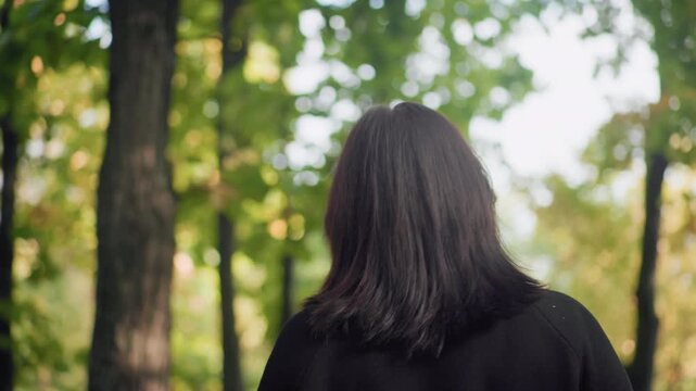 Medium back view of autumn lover strolling under leafy canopy, sipping warm drink with unhurried steps, black jacket and boots moving along forest path as sunlight filters through leaves