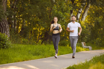 Fototapeta premium Smiling young man and woman jogging together along park path surrounded by green trees. Active couple enjoying morning run outdoors, maintaining healthy lifestyle, fitness routine and positive mood.