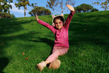 A girl wearing headphones and glasses is lying on the grass. cheerful child is playing and laughing in the park.