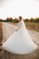 Bride in elegant white wedding dress standing on rural road at sunset. Romantic bridal portrait with soft golden light and natural outdoor background, symbolizing love, beauty, and serenity.