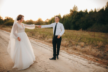 Bride and groom walking along a country road at sunset. The bride wears a beautiful white dress, the groom in a classic suit. Romantic wedding photo shoot outdoors with warm light and genuine emotions