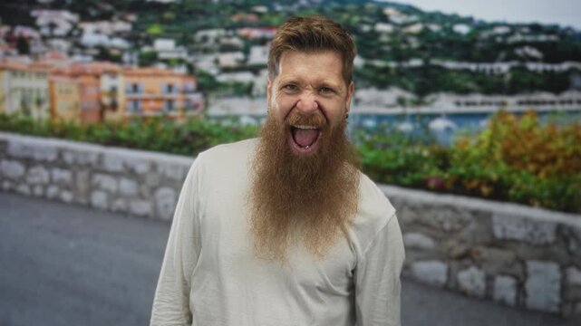 Man with long beard makes rock horns gesture with hand while sticking out tongue on beach promenade; spirited enthusiasm.