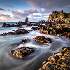 Coastal rocks bathed in soft light, with swirling water.  A dramatic, rocky shoreline meets the sea.  Clouds build in a dramatic sky