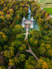 Aerial view of historic castle symmetrical twin towers and central hall framed by forest, moats, and tree-lined path, with open fields and residential zone beyond under warm sunrise sky.