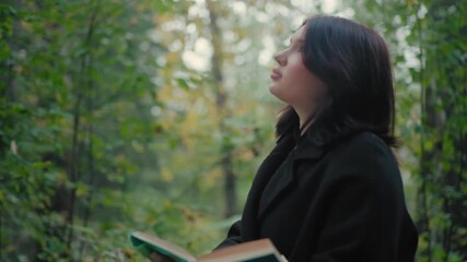 female tourist in autumn coat flips book pages while strolling forest path, glancing around, soft bokeh greenery, calm mood, quiet reading break during walk, peaceful outdoor escape under leaves