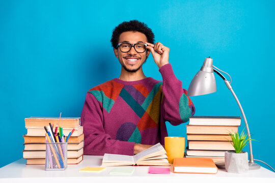 Funny stylish man with glasses studying at desk surrounded by books and supplies against bright blue background
