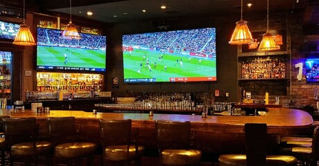 Sports bar interior shows multiple screens displaying soccer game. Wooden counter, stools, bottles visible. Warm lighting creates inviting atmosphere.