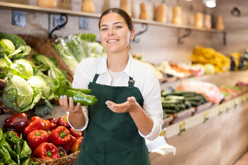 Successful young female owner of fruit and vegetable shop standing next to colorful produce stands, offering fresh green peppers..