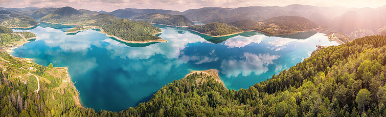 Zaovine lake reservoir is reflecting clouds, surrounded by rolling green hills, dense forests, and...