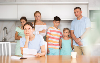 Fototapeta premium Upset mother reads and looks through financial documents. Sad wife with children in the background