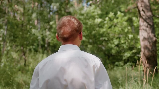 Close up back view of man in white shirt with glasses turned around in forest under daylight surrounded by green trees expressing excitement and curiosity in natural outdoor woodland environment