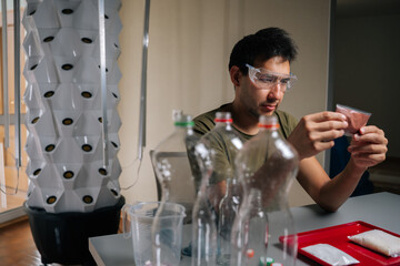 Agricultural technician wearing safety glasses examining fertilizer packet while preparing to nourish plants in vertical hydroponic tower, surrounded by measuring equipment, recycled plastic bottles.