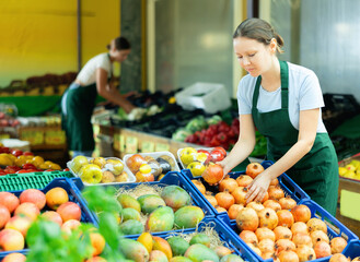 Positive skilled saleswoman in green apron working in fruit and vegetable section of supermarket,...