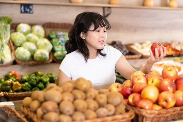Obraz premium Delighted young woman purchaser choosing red apples at the counter in grocery store