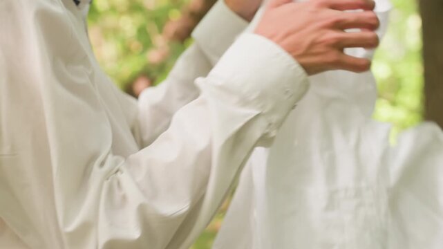 Closeup of young man putting on white coat with focus on sleeve and fabric detail, natural light filtering through surrounding forest greenery creating soft atmosphere of outdoor preparation