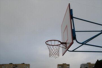 A basketball hoop and backboard are outside with buildings visible beneath a cloudy sky.
