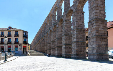 Segovia's massive Roman Aqueduct is a monumental wall of stone arches slicing through the city center. Its immense scale dominates the paved plaza and nearby urban buildings.