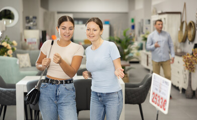 Fototapeta premium Interested positive female friends choosing functional furniture for homes in shop, chatting carefree while examining displayed samples..