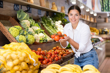 Interested smiling young girl shopping for organic vegetables in farm store, choosing ripe tomatoes from wicker box on produce display..